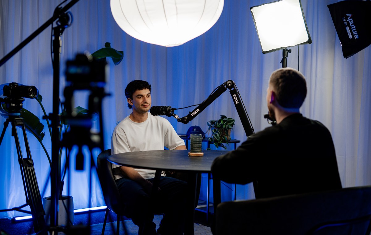 Bailey Williams seated at the studio table with a microphone in front of him, Jackson Morton with his back to camera, studio lights and cameras around them.