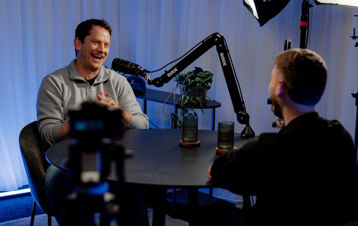 Stu Moerman and Jackson Morton seated across a dark table in the studio, microphones between them, a camera in the foreground.